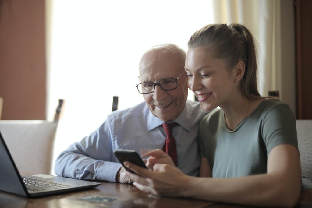 Young adult woman wearing casual shirt helps older man in formal shirt to use a mobile phone. They are sitting in a table that has a laptop on top.